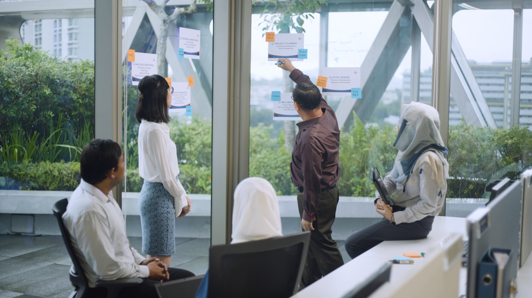Five people are in a modern office near a window with papers and sticky notes, one man is writing on the glass.